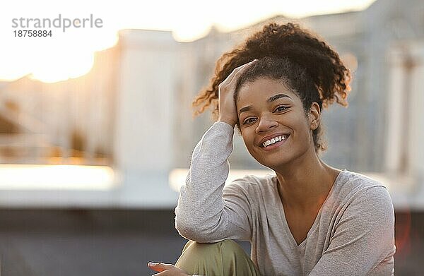 Junges glückliches afrikanisches amerikanisches Mädchen  das in die Kamera schaut  während es am sonnigen Morgen auf der Dachterrasse sitzt  lächelnde gemischtrassige Frau  die sich nach dem Training im Freien bei Sonnenaufgang entspannt. Glück und Wohlbefinden