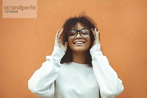 Fröhliches  positives  junges afroamerikanisches Mädchen mit Brille  lächelnd  afroamerikanische Frau  die eine Brille hält und mit den Händen das Gestell begradigt  breit lächelnd in die Kamera schaut  im Freien an einer orangefarbenen Wand stehend