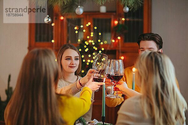 Fröhliche junge Freunde stoßen mit Gläsern Wein am weihnachtlich geschmückten Tisch zu Hause an