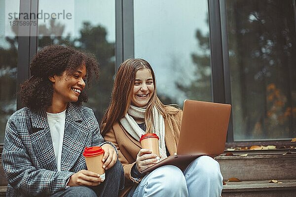 Zwei glückliche  positive  multiethnische Teenagermädchen mit Computer und trinken Kaffee zum Mitnehmen im Freien  schwarzweiße beste Freundinnen  die auf den Laptopbildschirm schauen und lachen  während sie sich im Herbstpark entspannen