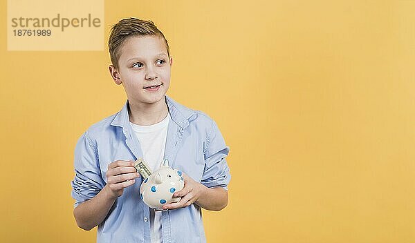 Lächelnder Junge  der Geldscheine in ein weißes Sparschwein aus Keramik einlegt