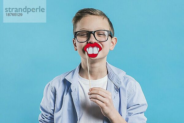 Junge mit schwarzer Brille hält lächelnd Requisite vor seinem Mund gegen blaün Hintergrund