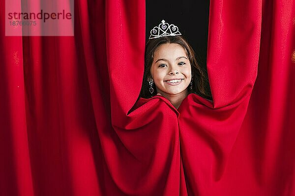 Lächelndes hübsches Mädchen mit Krone  das aus dem roten Vorhang schaut