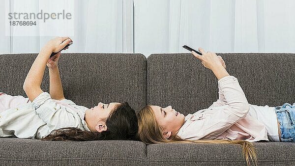 Glückliche Freundinnen liegen auf dem Sofa und benutzen ein Smartphone