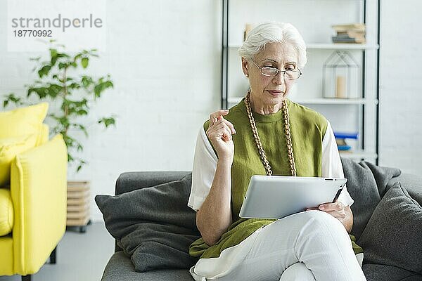Ältere Frau sitzt auf dem Sofa und schaut auf ein digitales Tablet