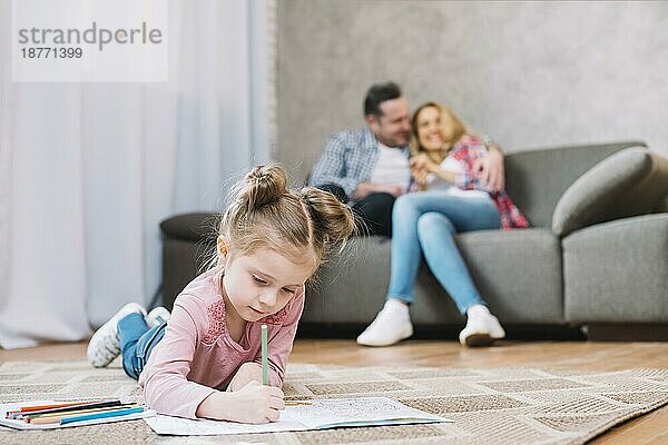 Kleines Mädchen Zeichnung Buch liegend Boden  während ihre liebevollen Eltern sitzen Sofa