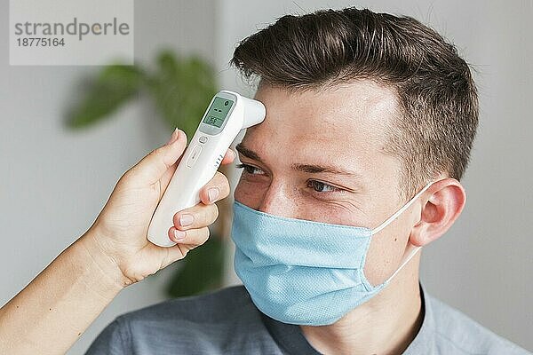 Mann mit medizinischer Maske  der mit einem Thermometer die Temperatur im Büro überprüft