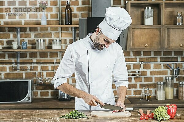 Glücklicher junger männlicher Koch schneidet roten Chili mit Messer Küchentisch. Foto mit hoher Auflösung