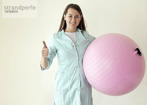 Lächelnde Physiotherapeutin  die einen Gymnastikball hält und Daumen nach oben zeigt