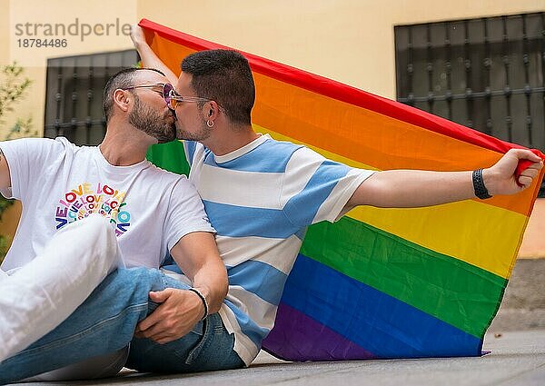 Porträt eines schwulen männlichen Paares  das auf dem Boden sitzt und sich mit einer Regenbogenfahne auf einer Pride Party küsst  lgbt Konzept