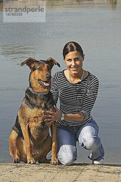 Junge Frau sitzt mit Dobermann-Schäferhund Mischling am Wasser