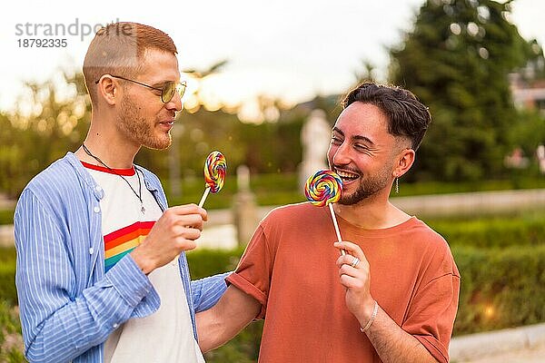 Porträt eines lächelnden Hochzeitspaares  das einen Lutscher im Park bei Sonnenuntergang in der Stadt isst. Vielfalt und lgbt Konzept