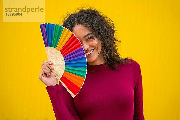 Porträt einer lateinischen Frau lächelnd mit einem Regenbogen lgbt Fan auf einem gelben Hintergrund