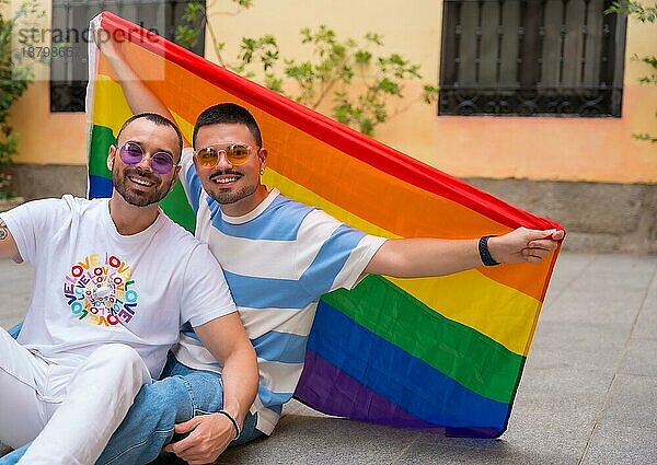 Porträt eines schwulen männlichen Paares auf dem Boden sitzend mit Regenbogenfahne bei einer Pride Party  lgbt Konzept