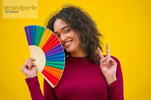 Porträt einer lateinischen Frau lächelnd mit einem Regenbogen lgbt Fan auf einem gelben Hintergrund  so dass der Sieg Symbol