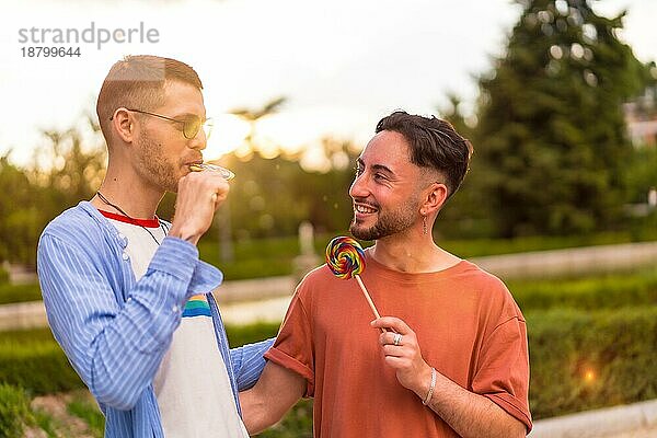 Porträt eines lächelnden Hochzeitspaares  das einen Lutscher im Park bei Sonnenuntergang in der Stadt isst. Vielfalt und lgbt Konzept