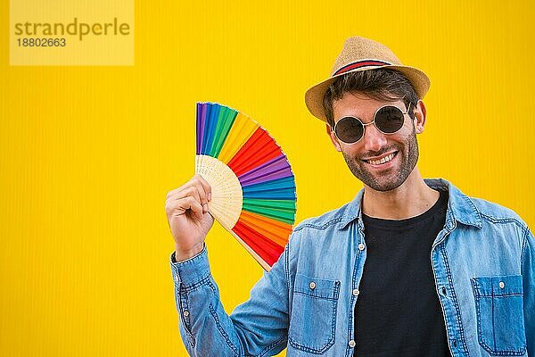 Porträt eines kaukasischen Mannes in einem Hut lächelnd mit einem Regenbogen lgbt Fan auf einem gelben Hintergrund