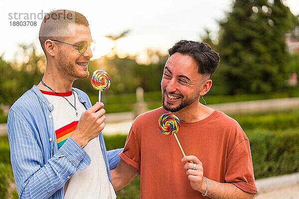 Porträt eines lächelnden Hochzeitspaares  das einen Lutscher im Park bei Sonnenuntergang in der Stadt isst. Vielfalt und lgbt Lebensstil Konzept