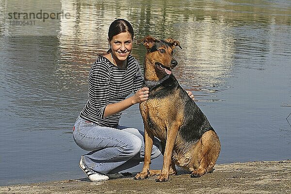 Junge Frau sitzt mit Dobermann-Schäferhund Mischling am Wasser