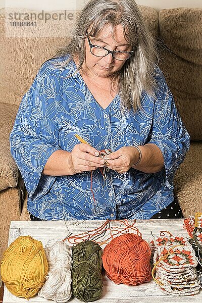 Vorderansicht einer älteren Frau mit weißem Haar und Brille  die mit ihrer Wolle im Vordergrund häkelt