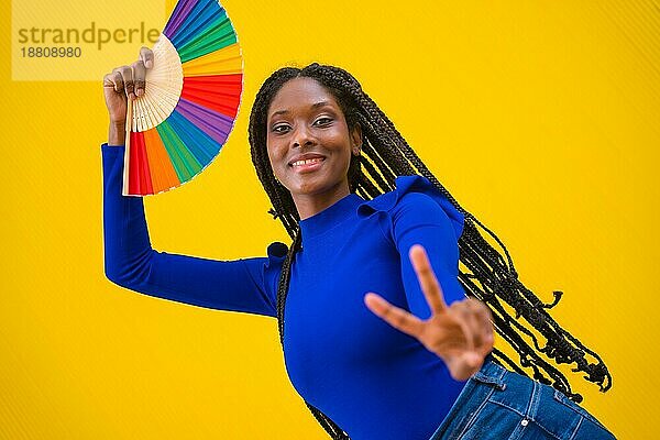 Porträt einer schwarzen ethnischen Frau tanzt mit einem Regenbogen lgbt Fan auf einem gelben Hintergrund