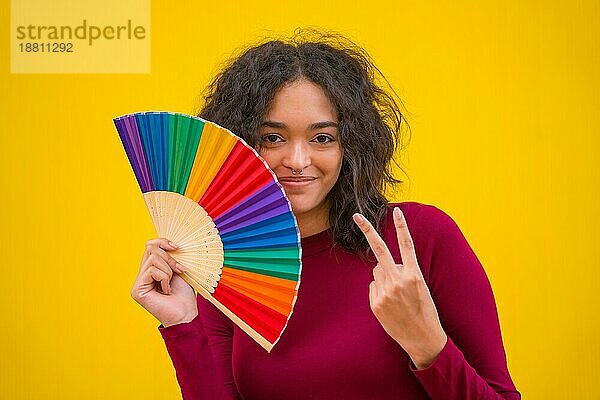 Porträt einer lateinischen Frau lächelnd mit einem Regenbogen lgbt Fan auf einem gelben Hintergrund  so dass der Sieg Symbol