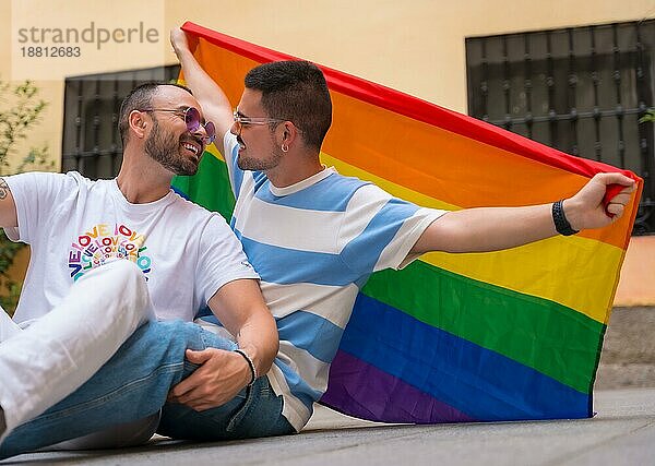 Porträt eines schwulen männlichen Paares auf dem Boden sitzend mit Regenbogenfahne bei einer Pride Party  lgbt Konzept