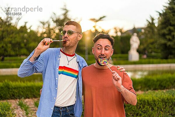 Porträt des lachenden Hochzeitspaares beim Essen eines Lutschers im Park bei Sonnenuntergang in der Stadt. Vielfalt und lgbt Konzept