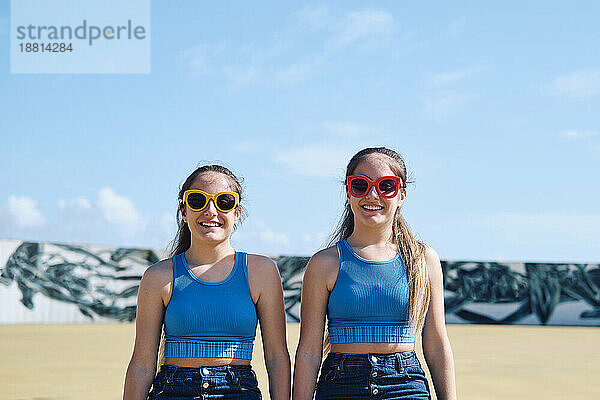 Lächelnde Zwillingsschwestern mit Sonnenbrille stehen an einem sonnigen Tag