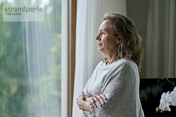 Nachdenkliche ältere Frau steht mit verschränkten Armen und schaut zu Hause durch das Fenster
