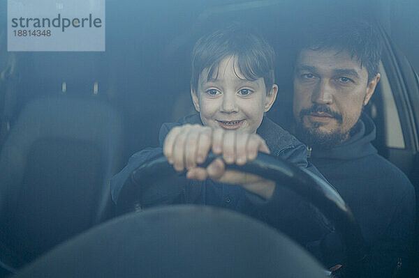Lächelnder Sohn lernt Autofahren  während sein Vater durch die Windschutzscheibe gesehen wird