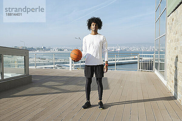 Junger Athlet mit Basketball steht an einem sonnigen Tag auf dem Pier