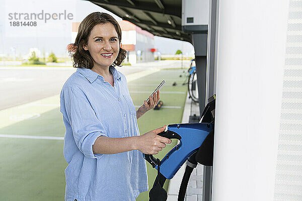 Lächelnde Frau hält Autoladegerät und Smartphone am Bahnhof