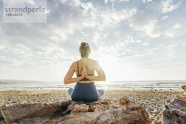 Frau praktiziert Yoga mit hinter dem Rücken verschränkten Händen und sitzt auf einem Felsen am Strand