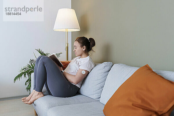 Woman with diabetes reading book sitting on couch at home