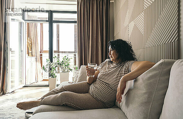 Nachdenkliche Frau mit einem Glas Wasser sitzt auf dem Sofa