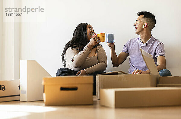 Lächelndes Paar genießt Kaffee und sitzt neben Pappkartons im neuen Zuhause