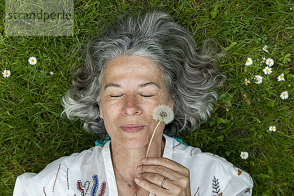 Ältere Frau liegt auf grünem Gras und hält Löwenzahn-Pusteball