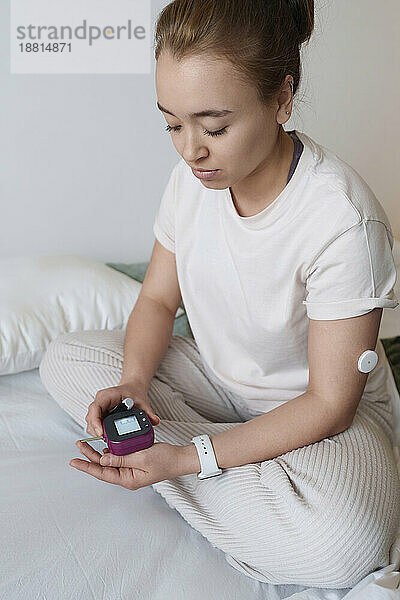 Frau mit Diabetes sitzt auf dem Bett und hält zu Hause ein Blutzuckermessgerät in der Hand