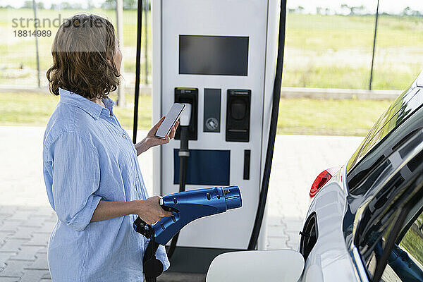 Frau hält Autoladegerät und Smartphone neben Elektroauto am Bahnhof