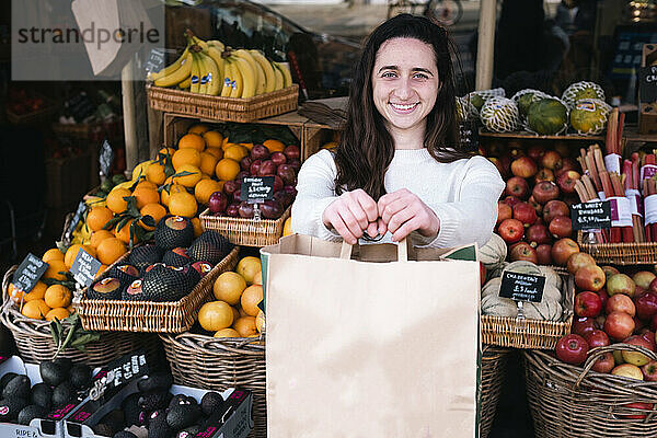 Glücklicher junger Lebensmittelhändler steht mit Papiertüte im Gemüseladen