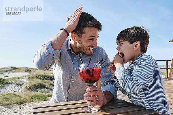 Fröhlicher Vater hält ein Glas Saft in der Hand  während sein Sohn am Tisch isst