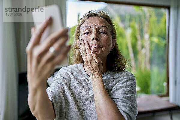 Ältere Frau schaut zu Hause auf ihr Smartphone und berührt ihr Gesicht
