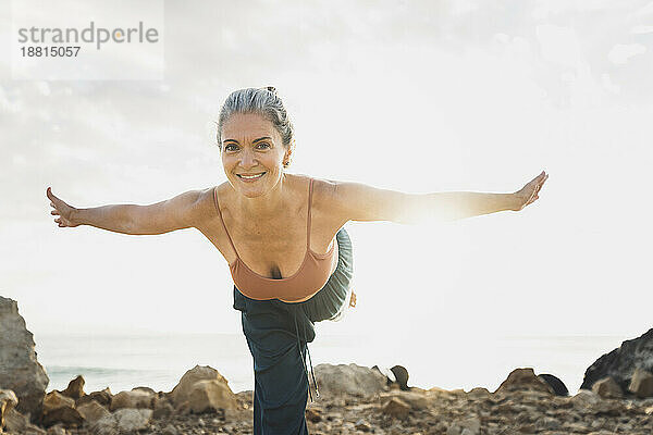 Lächelnde reife Frau praktiziert Yoga mit ausgestreckten Armen am Strand