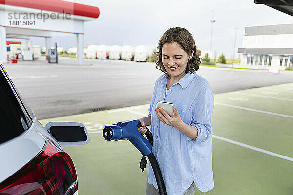 Lächelnde Frau benutzt Smartphone neben Elektroauto am Bahnhof