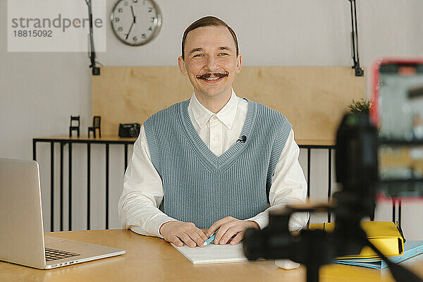 Lächelnder Mann sitzt im Klassenzimmer und blickt auf die Kamera  die den Online-Unterricht aufzeichnet