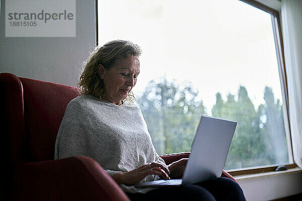 Ältere Frau sitzt zu Hause im Sessel und benutzt Laptop