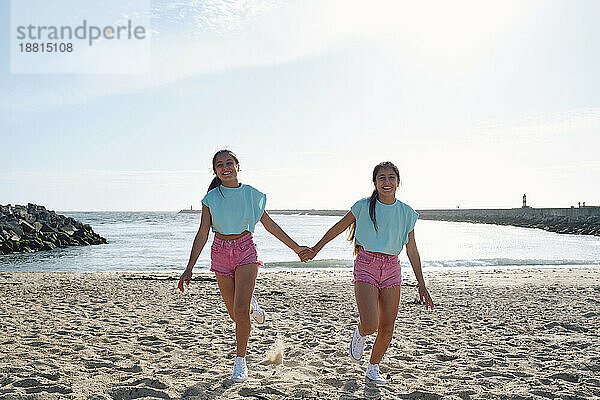 Sorglose Zwillingsschwestern halten Händchen und gehen am Strand spazieren