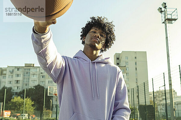 Selbstbewusster Athlet mit lila Kapuzenpullover hält Basketball auf dem Sportplatz