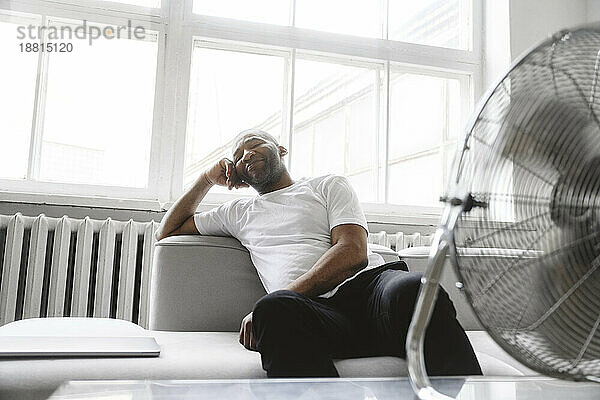 Mann entspannt sich zu Hause auf Sofa vor elektrischem Ventilator
